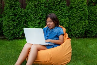 Girl Child Sits On A Pillow With An Open Laptop On The Green Grass In The Courtyard Of The Girl Child Sits On A Pillow With An Open Laptop On The Green Grass In The Courtyard Of The