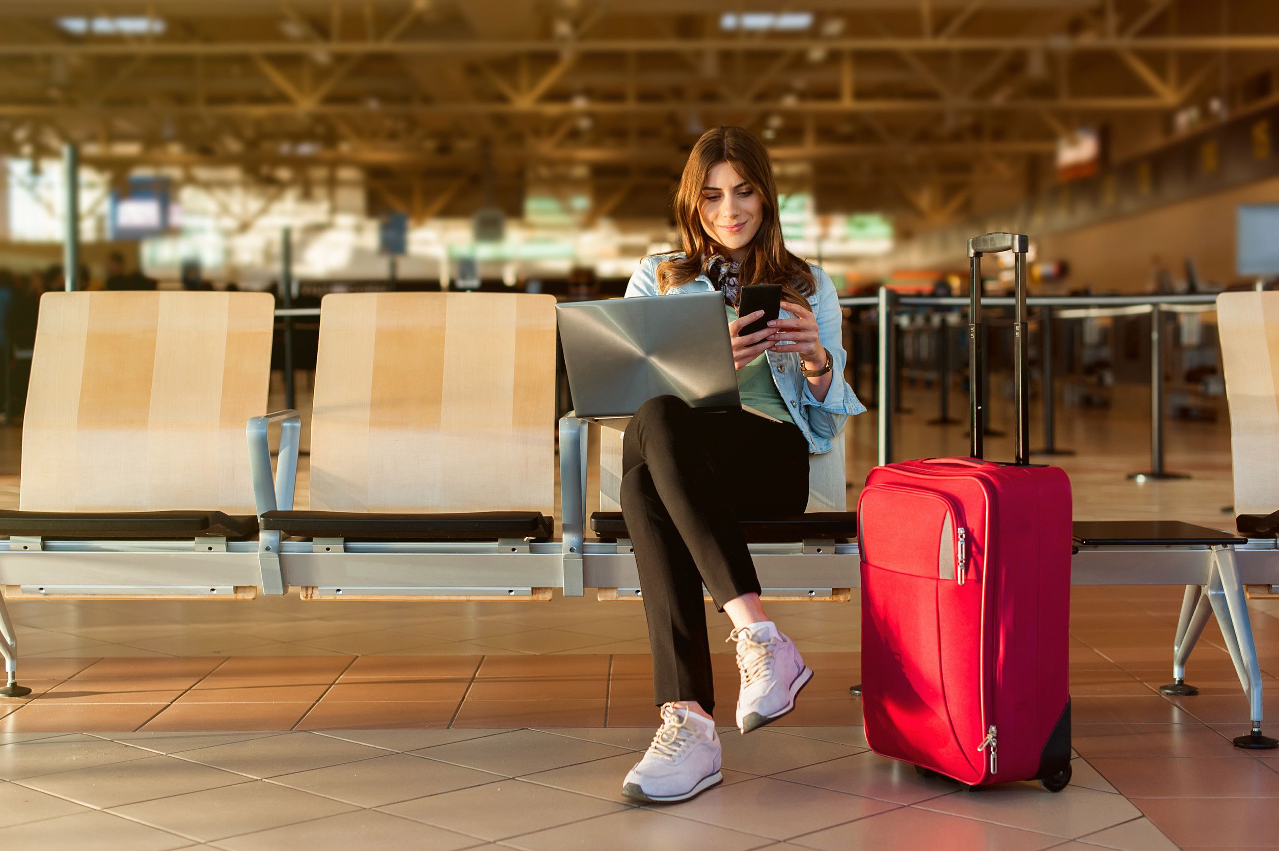 Jeune passagère à l'aéroport, assise dans le hall du terminal, utilisant son smartphone et son ordinateur portable. Jeune passagère à l'aéroport, assise dans le hall du terminal, utilisant son smartphone et son ordinateur portable.