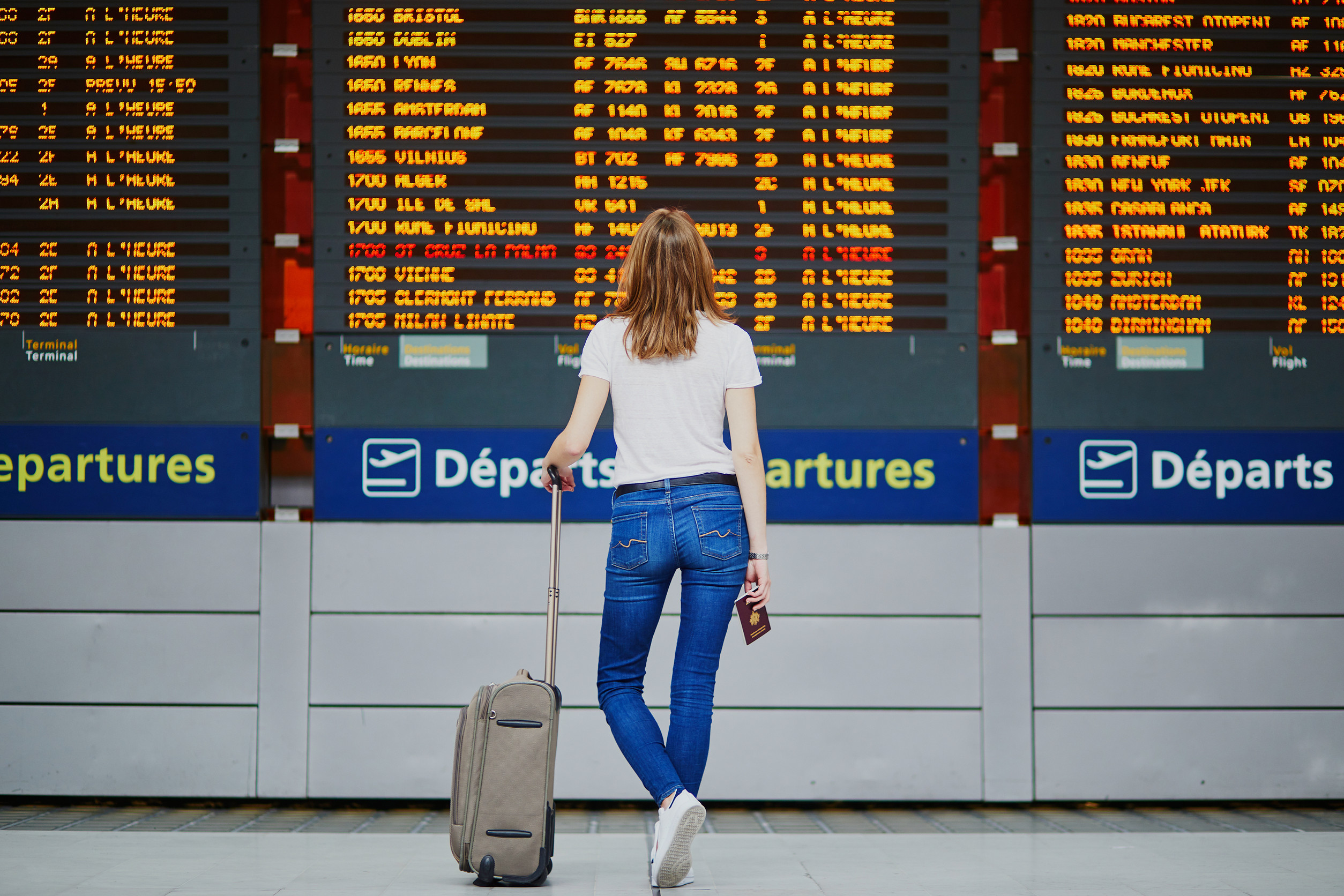 Mujer joven en el aeropuerto internacional con equipaje y pasaporte cerca de la pantalla de información del vuelo Mujer joven en el aeropuerto internacional con equipaje y pasaporte cerca de la pantalla de información del vuelo